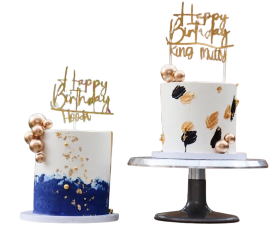 Two-tier modern birthday cake with gold and blue decorations and custom gold "Happy Birthday" toppers on a silver cake stand and glass table with a wooden fence and flowers in the background.