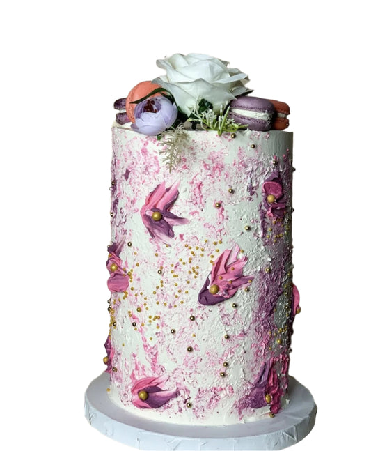 Tall floral-themed celebration cake with textured pink and white buttercream, decorated with hand-piped flowers, gold pearls, fresh white rose, and pastel macarons on top.