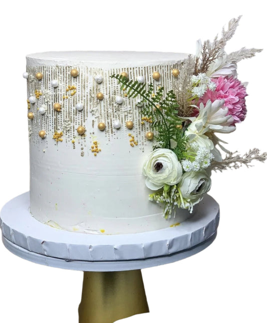 White buttercream cake with gold drip detailing, pearl-like accents, and fresh flower arrangement featuring ranunculus and greenery on the side.